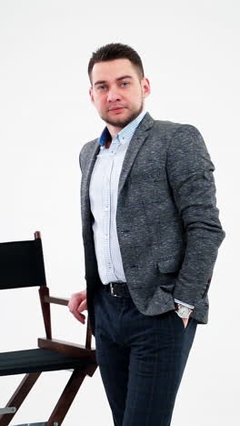 portrait of a young businessman in light studio. handsome man in elegant costume standing near the chair and looking at camera. vertical video