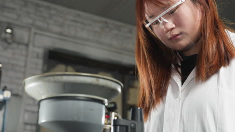 female engineer examines sample through microscope, hair cascading over shoulder, adjusts back, and taps on tablet in mechanical shop with industrial tools and machinery in background