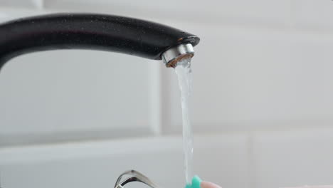 primer plano de un grifo en funcionamiento con una persona lavando el batedor usando una esponja verde bajo el agua que fluye en la cocina, salpicaduras de agua alrededor del batedor mientras se limpia, configuración moderna de la cocina
