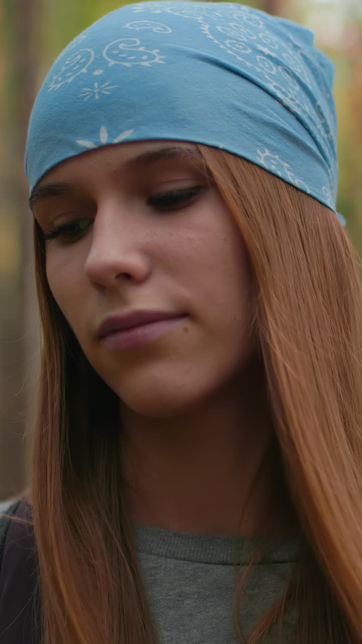 mujer con bandana azul caminando por el sendero del bosque con una sonrisa sutil, emanando confianza tranquila, mientras su amiga con camisa verde mira a su alrededor pacíficamente en un fondo borroso suave en medio de árboles altos