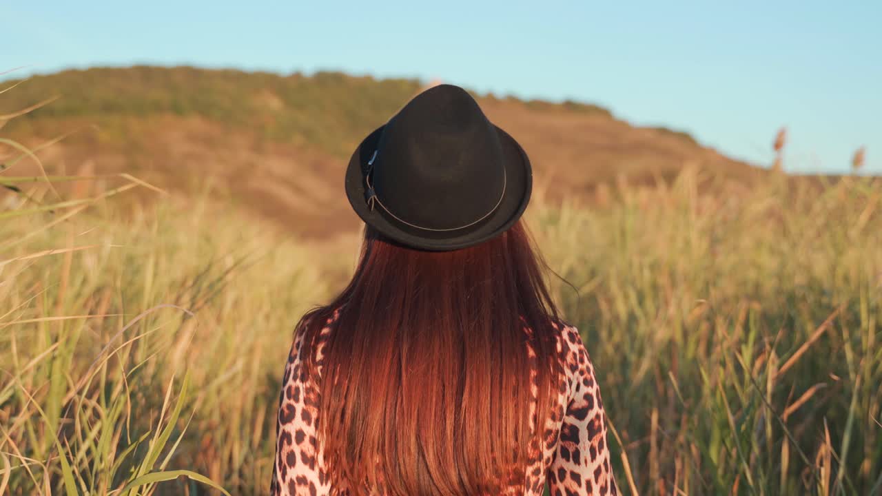 Young Caucasian woman wearing black hat, takes off hat and turns looking at camera smiling as she walks in rural tall green grass field, handheld close up