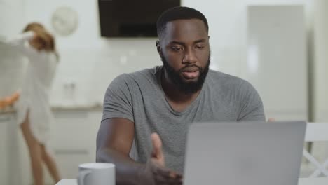 Un-Hombre-Negro-Triste-Trabajando-Con-Una-Computadora-Portátil-En-La-Cocina-Abierta.-Un-Joven-Loco-Escribiendo-Un-Cuaderno