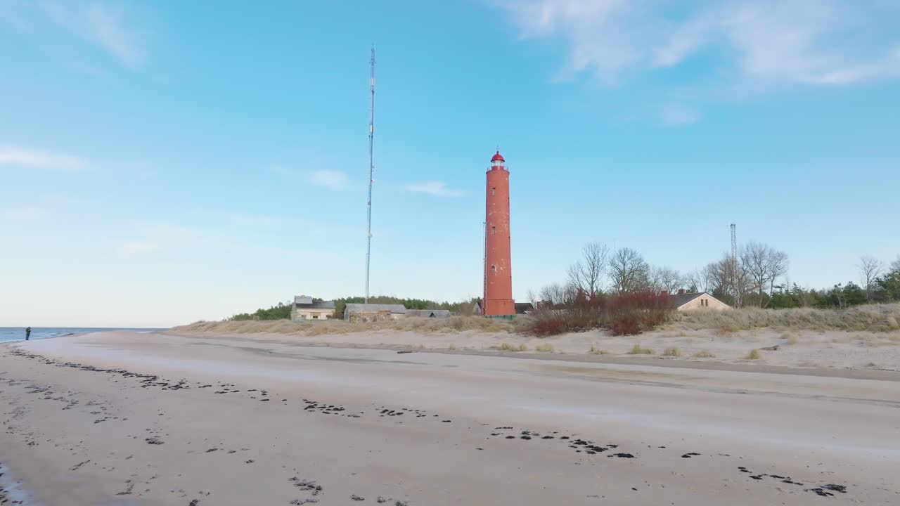 vista aérea del faro akmenrags de color rojo, costa del mar báltico, letonia, playa de arena blanca, mar en calma, día soleado con nubes, tiro de drones anchos y bajos avanzando