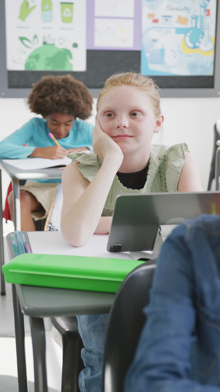 Vertical video of happy caucasian schoolgirl at desk with tablet, listening in class, copy space