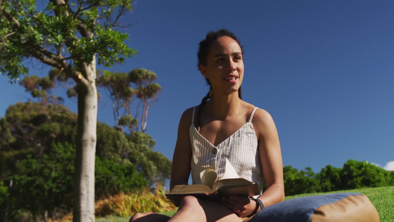 African american woman sitting on grass reading book in park