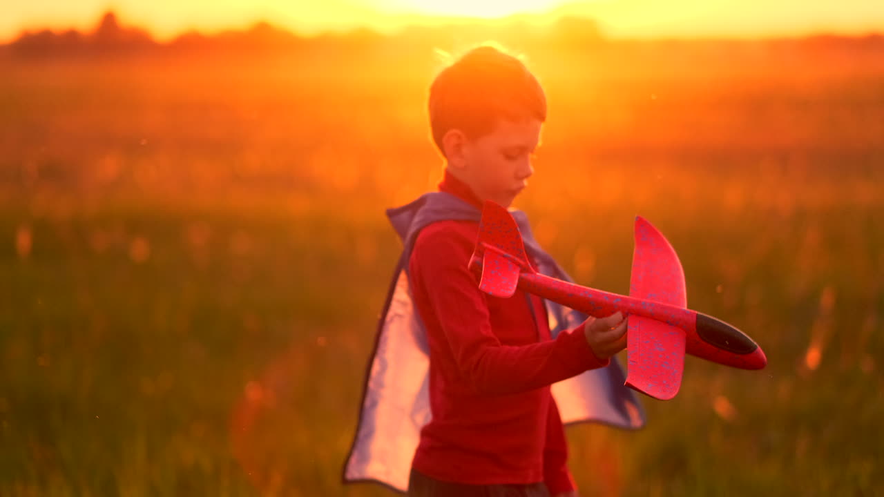 el niño con el disfraz de un superhéroe corriendo en una capa roja riendo al atardecer en el campo de verano representando que él era el piloto del avión