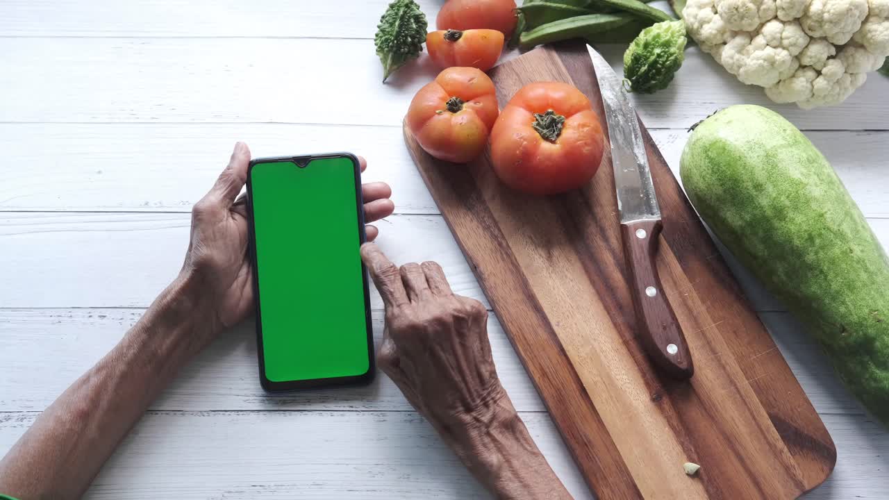 persona de edad avanzada que utiliza un teléfono inteligente mientras prepara verduras