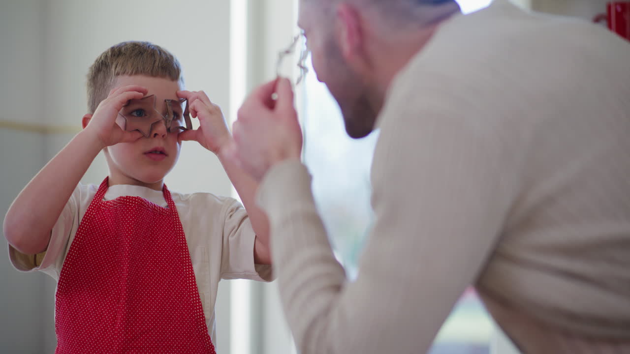 un niño hace reír a su padre con cortadores de galletas en la cocina.