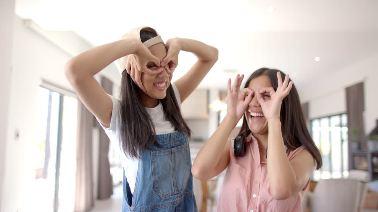 Biracial girls form heart symbols with hands over eyes, laughing at home