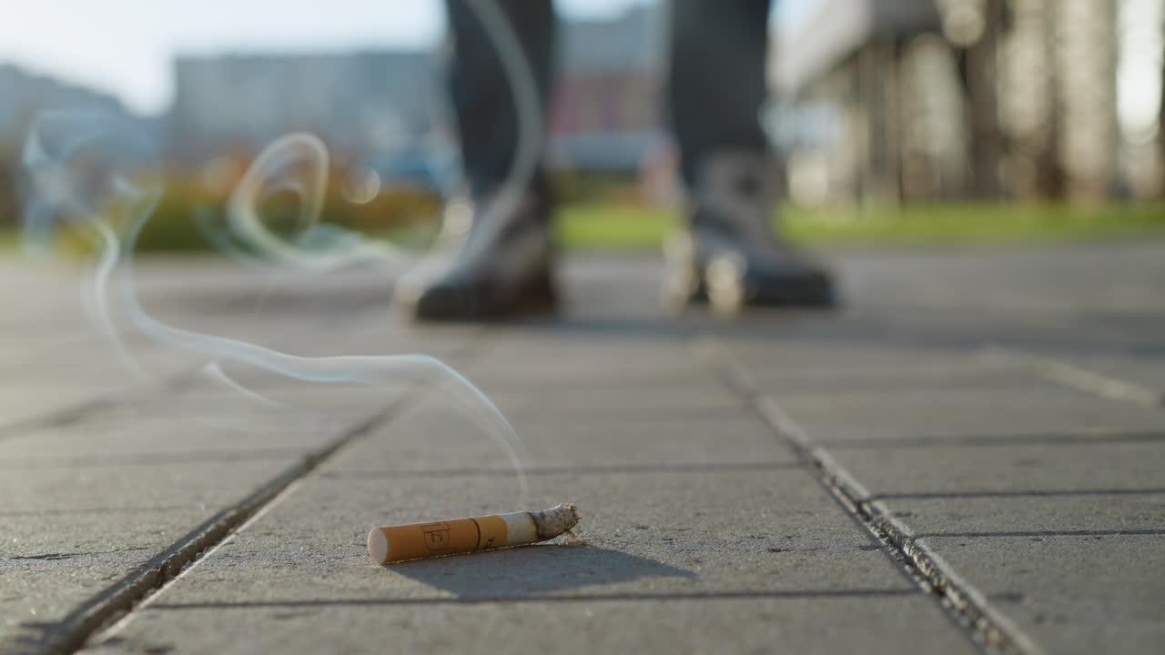 close up of burning cigarette on pavement with swirling smoke rising into air and blurred leg view of person standing nearby in outdoor urban environment under daylight