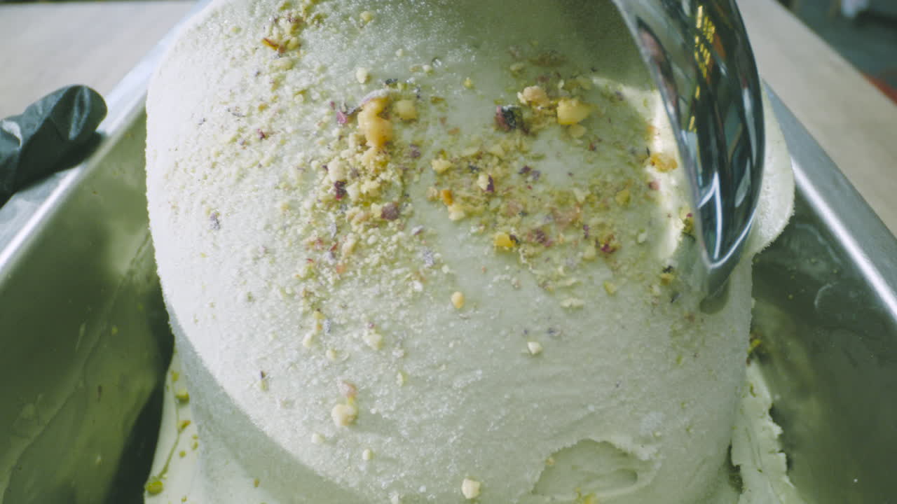 A close-up of creamy pistachio ice cream, with visible pieces of the nut, being scooped out with a metal scoop.
