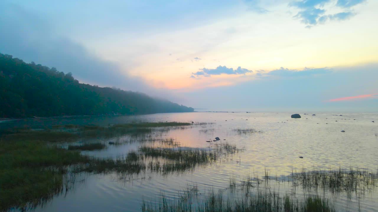 Stunning aerial drone footage gliding over grassy and reed filled beach area and continiues to fly over Baltic ocea sea during orange sunset, steep cliff bank in fog and mist visible on the left