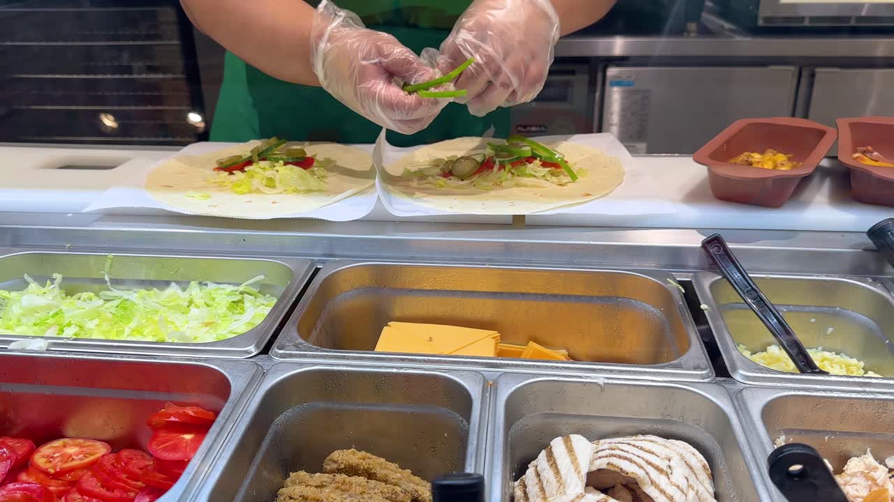 Un lavoratore che indossa guanti di plastica aggiunge peperoni verdi a delle tortillas a un bancone alimentare.