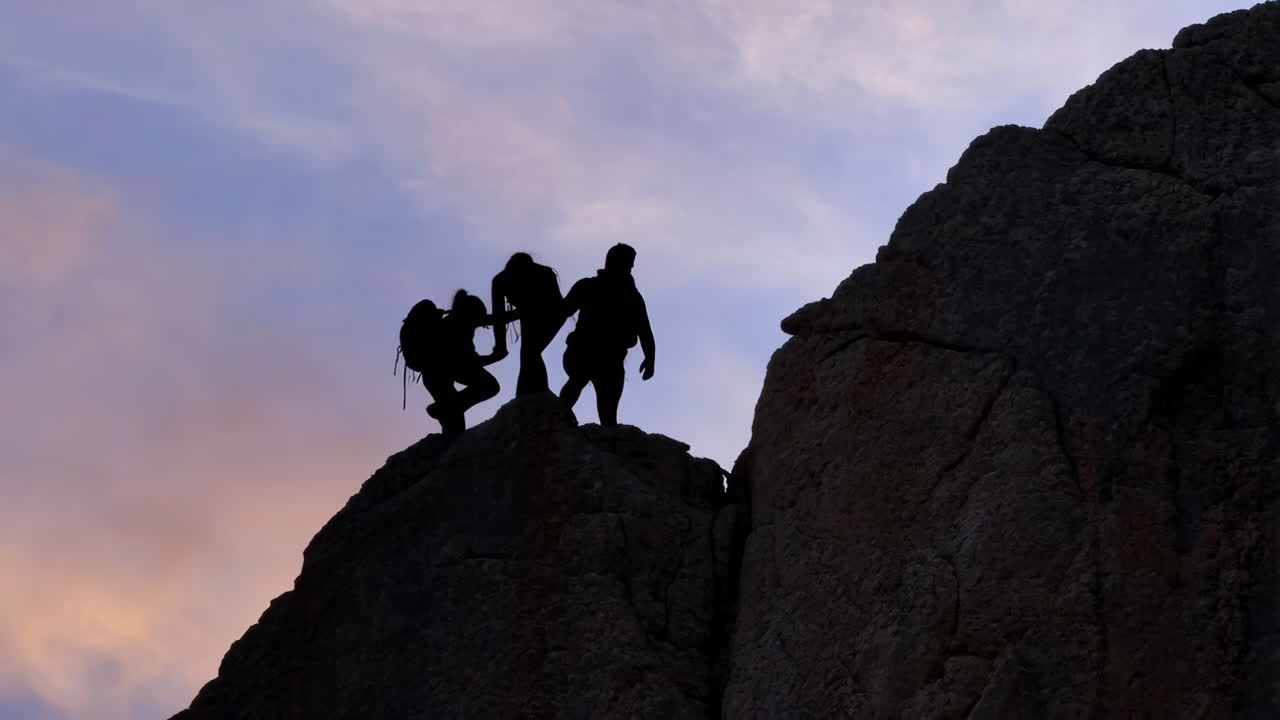 Silhouettes of two people assisting each other while climbing a rocky peak at sunset