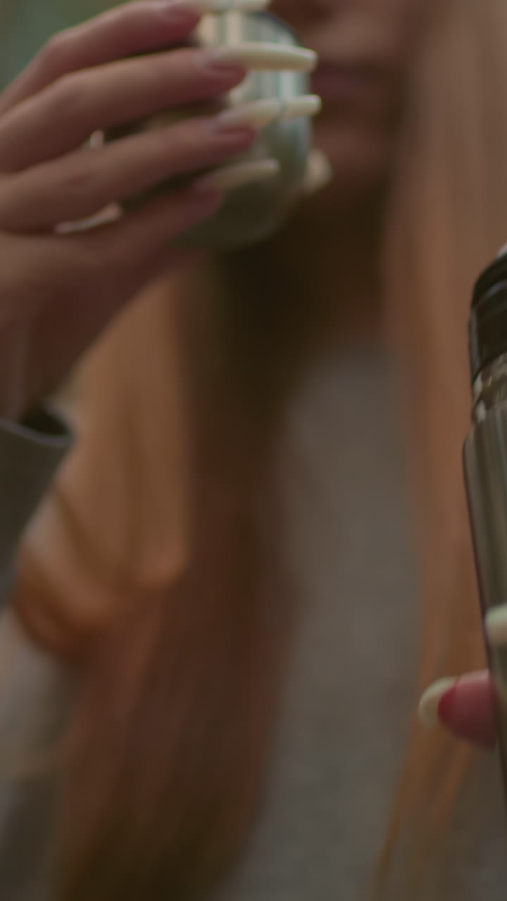 Close-up of young woman pouring water from thermos into its cap in peaceful forest setting, capturing her well-manicured nails drinks from it amidst blurred greenery background