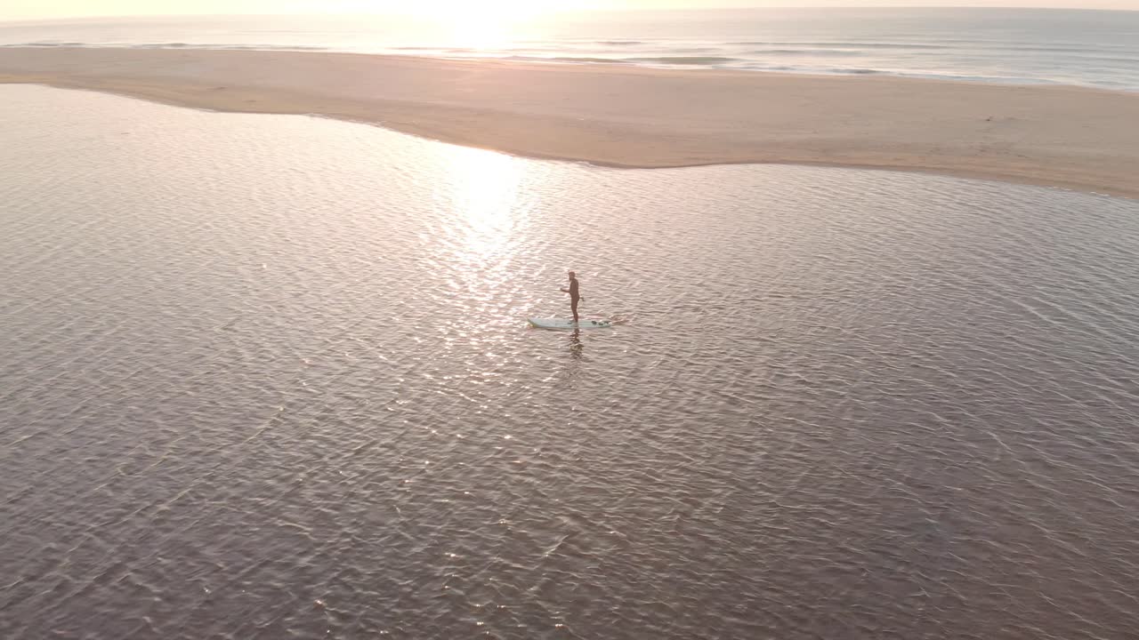 Ripresa aerea che orbita attorno a un uomo su una tavola da stand up paddle su un lago vicino all'oceano, con il sole che sorge sopra l'orizzonte sulla costa dell'Australia meridionale.