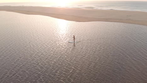 aerial shot orbiting around a man on a stand up paddle board on a lake near the ocean with the sun rising above the horizon on the coast in southern australia