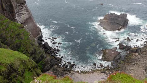 A-slow-tilting-shot,-revealing-a-rocky-bay-with-waves-crashing-against-rocks-and-a-tall-sea-cliff-in-the-ocean-while-seabirds-fly-around-the-cliffs-of-a-seabird-colony,-Handa-Island,-Scotland