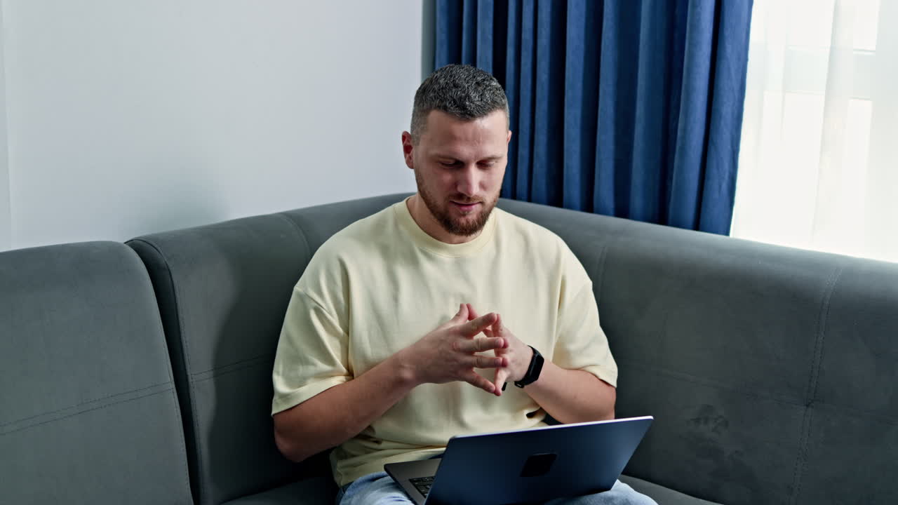 Man sits on couch using laptop in a bright living room with curtains drawn during daytime hours in an urban setting