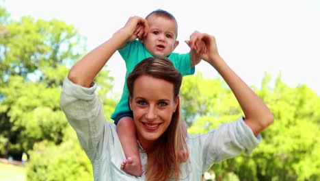 Happy-mother-holding-cute-baby-son-on-her-shoulders-in-the-park