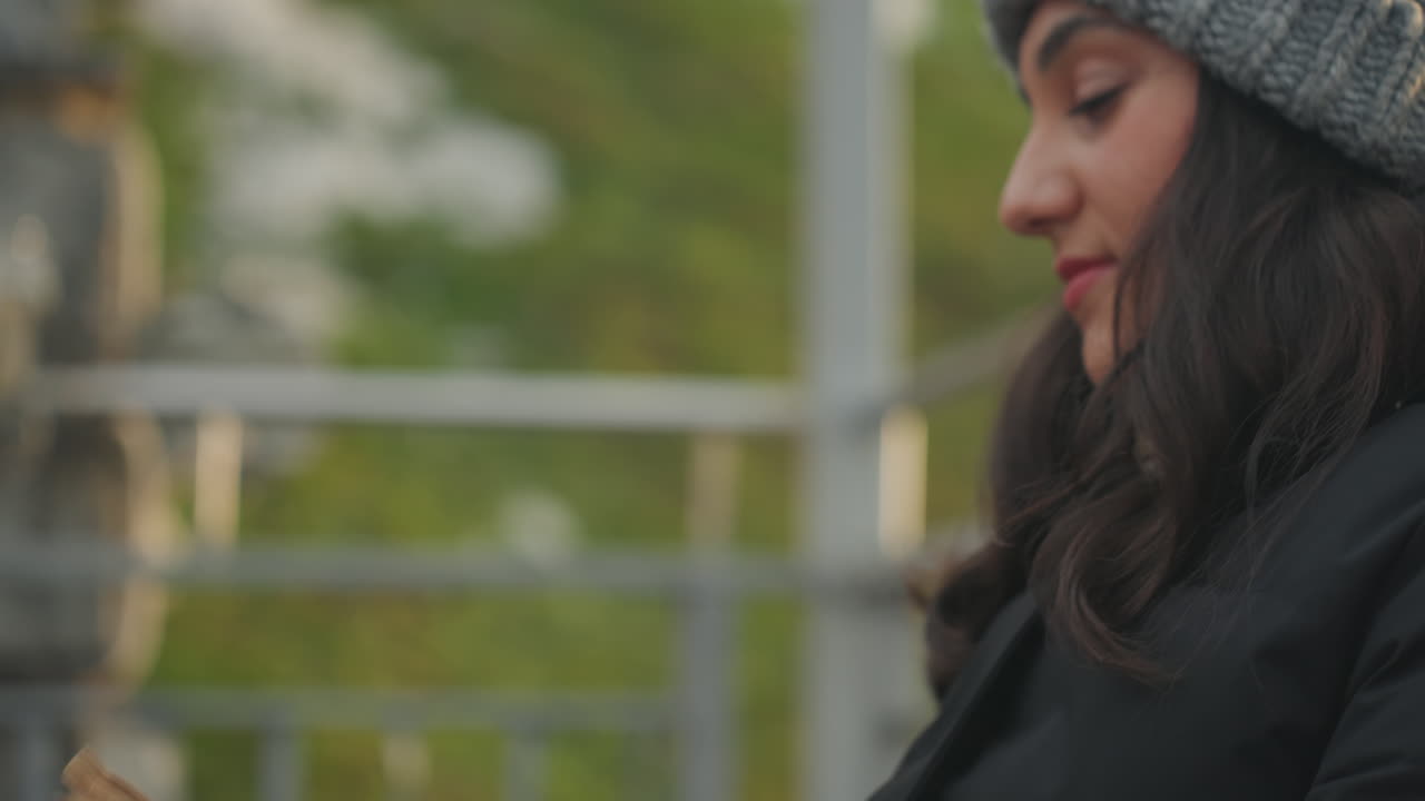 Giovane donna che sfoglia pagine su una panchina al crepuscolo, sorriso delicato, berretto di lana e giacca calda, primo piano di mani e viso, coinvolgimento narrativo e atmosfera rilassata, ambiente tranquillo del parco con sottile ringhiera sullo sfondo.