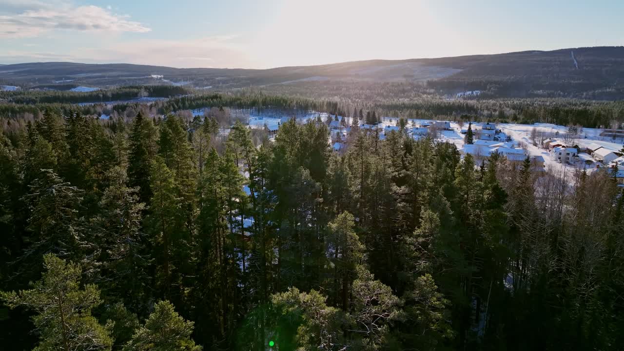 Flygfoto över vinterklädd skog med en glimt av byn Skorped, Sverige, där solsken tränger igenom träden