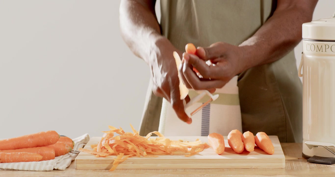 Uomo afroamericano con grembiule a righe che raccoglie una carota, pelandola con un pelapatate per la preparazione del pasto.