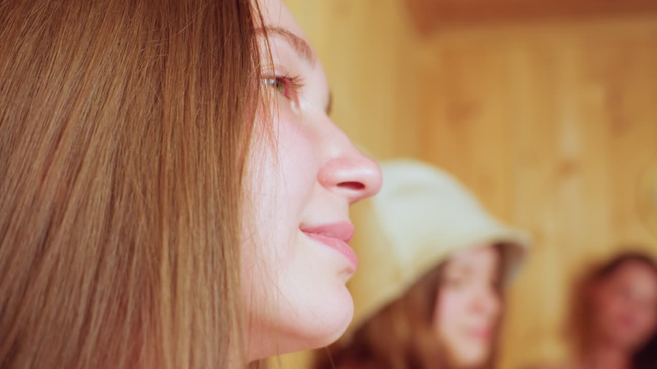 Inside wooden sauna, young adult woman wearing sauna hat looks thoughtful while two friends seated nearby raise glass mugs of juice in cheerful toast, sharing calm relaxed moment