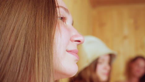 inside wooden sauna, young adult woman wearing sauna hat looks thoughtful while two friends seated nearby raise glass mugs of juice in cheerful toast, sharing calm relaxed moment