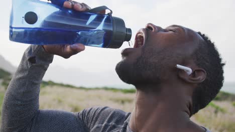 Hombre-Afroamericano-Bebiendo-Agua-De-Una-Botella-Mientras-Camina-Por-Las-Montañas