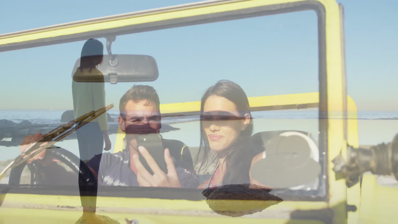 Taking selfie in yellow convertible, couple with surfboard silhouette in background
