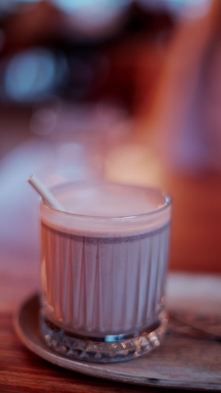 Close up of a hot chocolate in a glass with a straw on a table at a cafe. Vertical