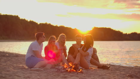 dos hombres y tres jóvenes estudiantes están sentados alrededor de una fogata en la playa con cerveza. una de las estudiantes está contando una historia interesante. otra está revisando su teléfono móvil al atardecer en la noche de verano en la costa del lago.