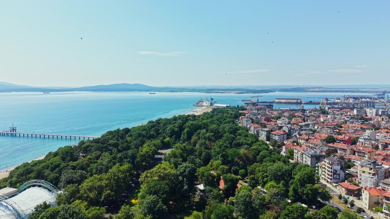 Berrak mavi gökyüzünün altında, sahil parkı, ağaçlarla çevrili gezinti yolu, liman, kiremit çatılı binalar ve uzaktaki adaları barındıran bir sahil şehrinin geniş açılı havadan görünümü.