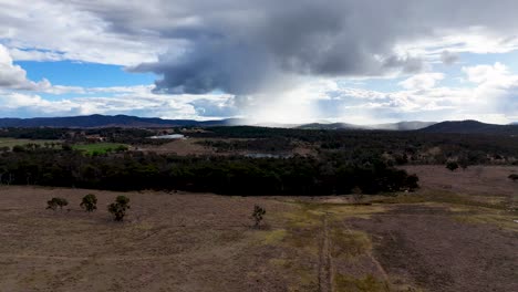 a drone ascends over a dry, open field with scattered trees near stanthorpe, australia, revealing distant forests, hills, and dramatic cloudy skies