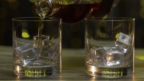 Pouring-of-whiskey,-cognac-or-brandy-from-bottle-into-glasses-with-ice-cubes.-Shiny-background