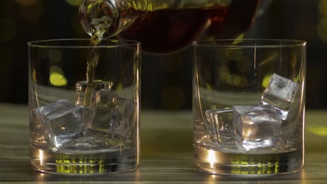 pouring of whiskey, cognac or brandy from bottle into glasses with ice cubes. shiny background