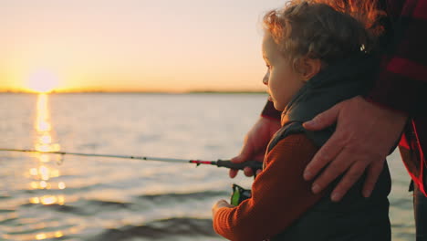 cute little boy is fishing by rod father or grandpa is helping to child happy family on river shore in sunset