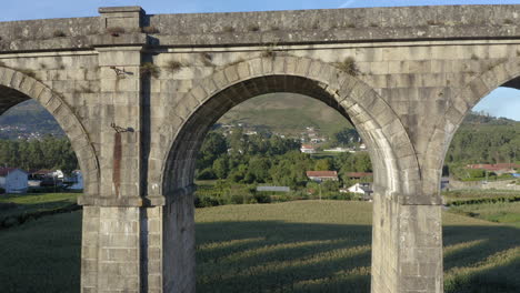 Aerial-Slow-Fly-Through-The-Arch-of-Old-Railway-bridge-surrounded-by-crop-fields-revealing-beautiful-Hills---Ponte-Seca,-Durrães,-Barcelos