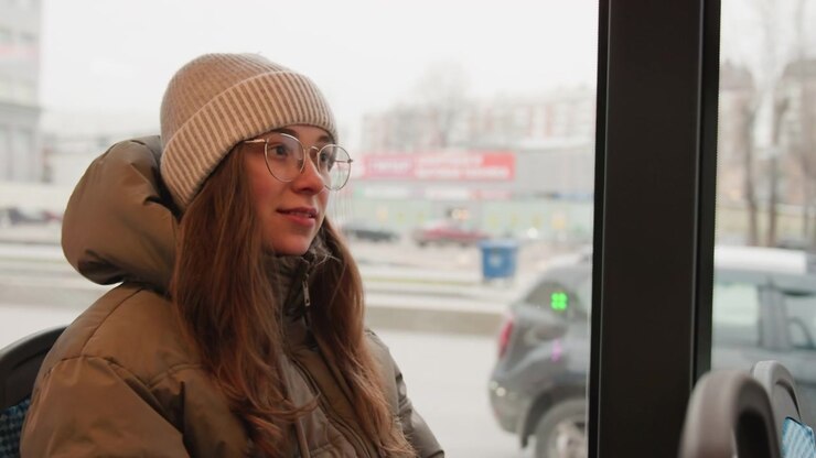 White woman on city bus, wearing beanie and puffer coat, looking through window at traffic, adjusting hair with reflective gaze, daytime urban commute, candid relaxed routine and people watching