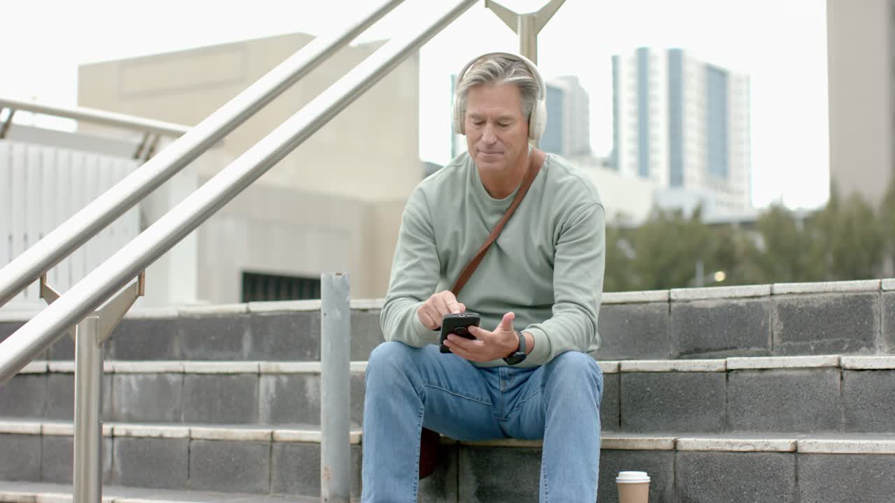 Man scrolling smartphone on plaza steps, hearing headphone audio, glancing at buildings