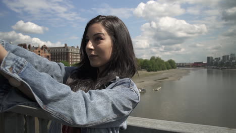 Attractive-and-smiling-Latina-tourist-taking-a-selfie-while-standing-on-the-railing-of-a-bridge-in-London,-slow-motion-shot