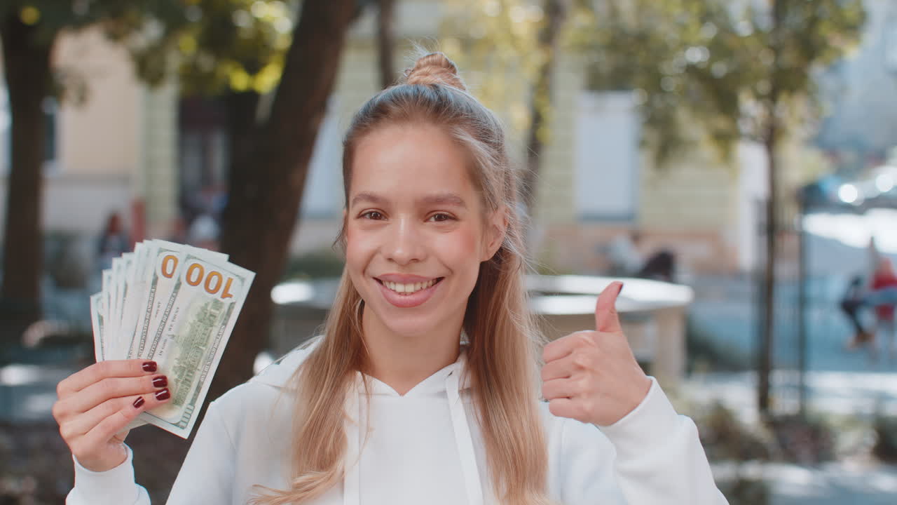 Felice giovane donna caucasica che tiene in mano un ventaglio di dollari e mostra il pollice all'insù per strada.