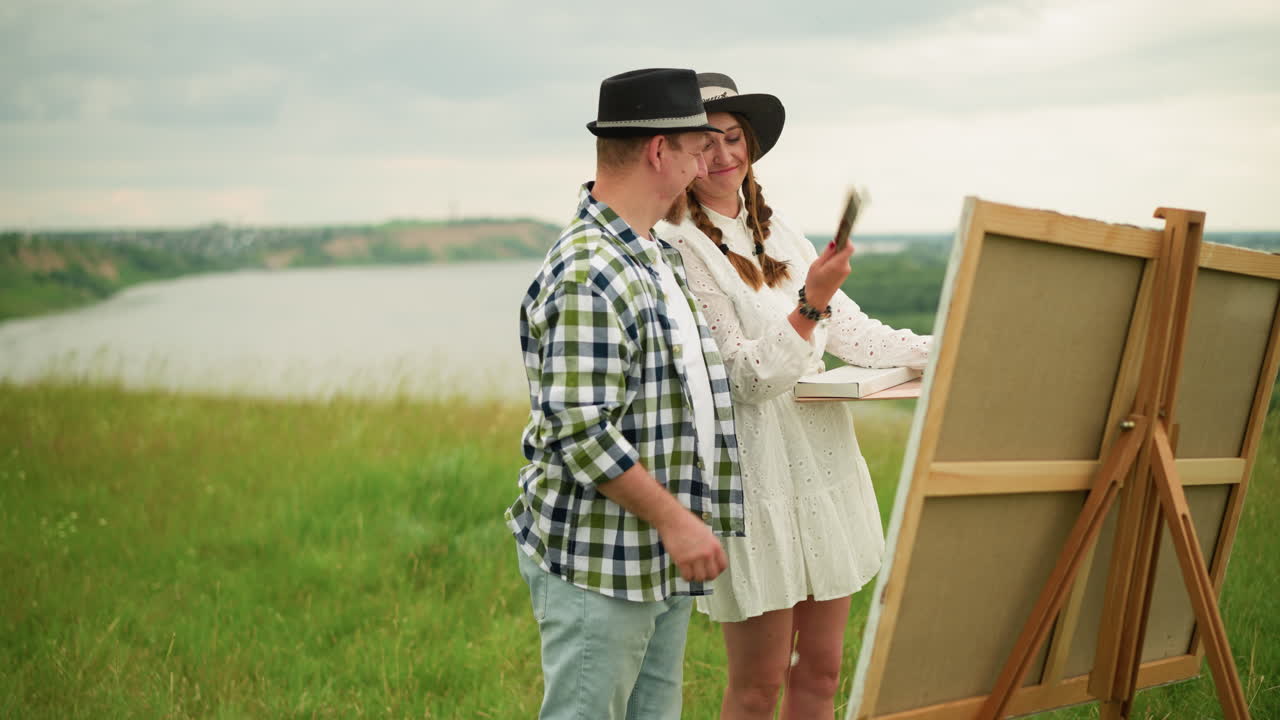 Two lovers stand in a green field, smiling at each other. The man, in a black hat and plaid shirt, and his partner in a white dress, share a moment of connection as they look at a board on an easel