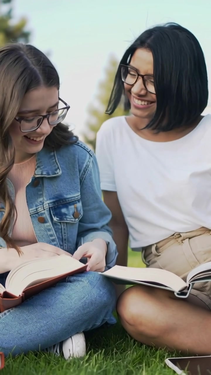 Video vertical: Pasando páginas sobre la hierba; una persona en vaqueros a la izquierda y otra en camiseta a la derecha se inclinan para estudiar libros y una tableta.