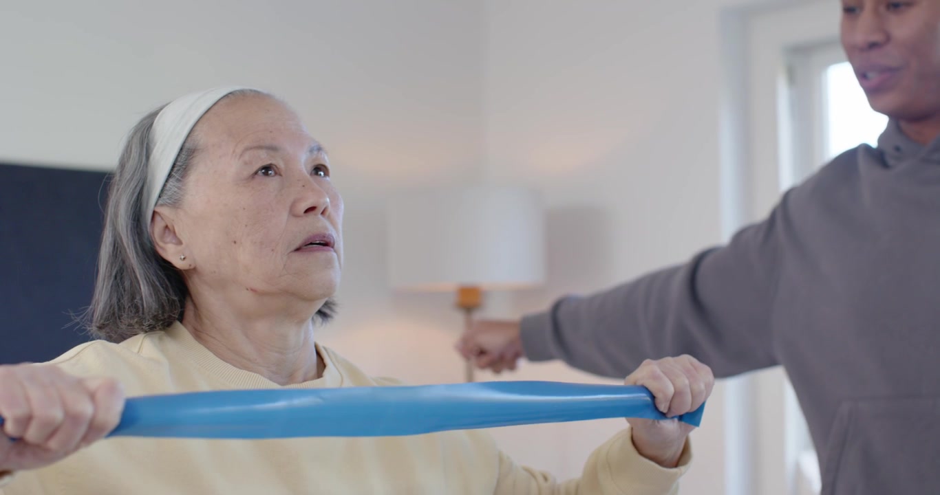 Senior Asian woman and African American caregiver placing hands pulling blue band at home for rehab
