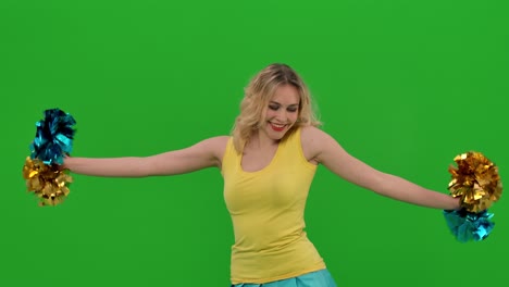 a cheerleader dances a jubilant dance with pompoms, gracefully crouches, does a vertical split. a blonde in a yellow and blue uniform is training in the studio on a green screen. close up. slow motion