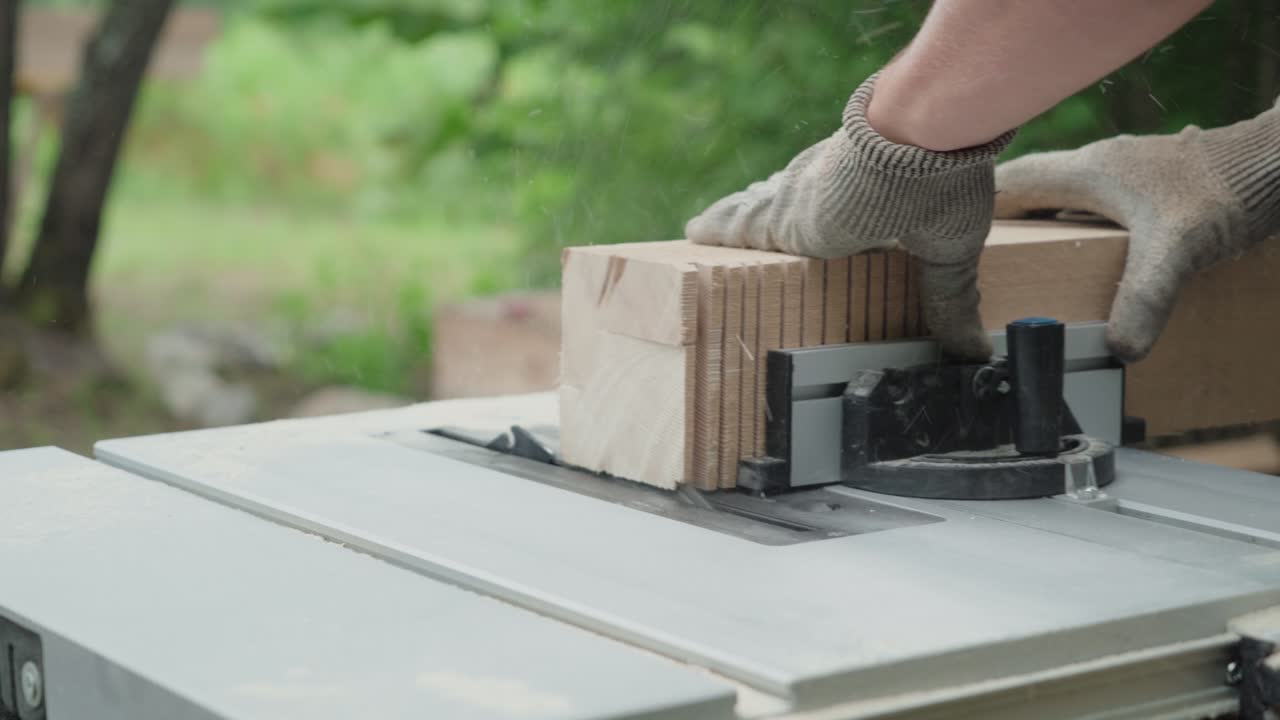 Primo piano di mani guantate che spingono legno impilato attraverso una sega circolare da banco per tagli precisi all'esterno