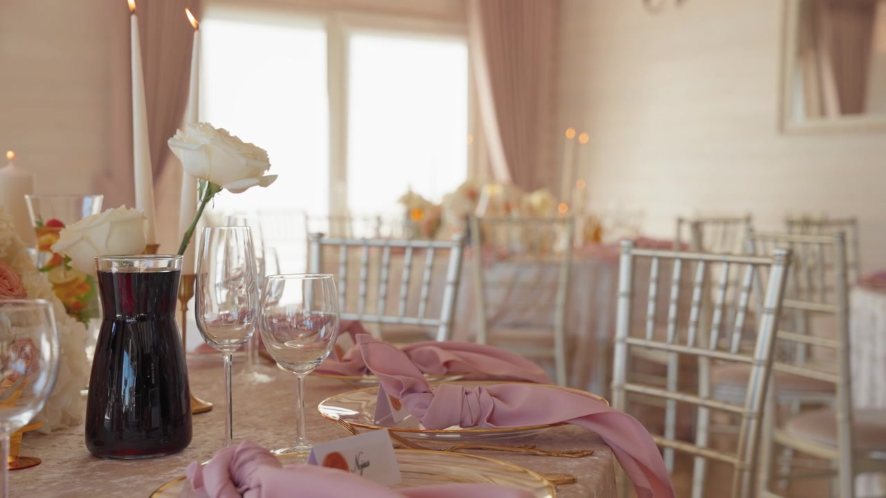 Beautifully arranged wedding table features pink napkins, gold plates, glassware, white roses, candles, and natural light. Delicate, airy mood highlights luxury event venue in close-up shot.