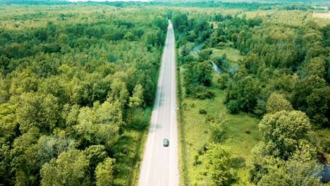 wide shot aerial drone footage tracking a sedan car cruising on an empty road surrounded by a beautiful large forest and river